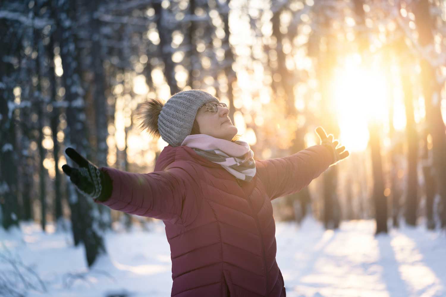 A person in winter gear standing with arms outstretched with the sun shining through the tree trunks in the background.