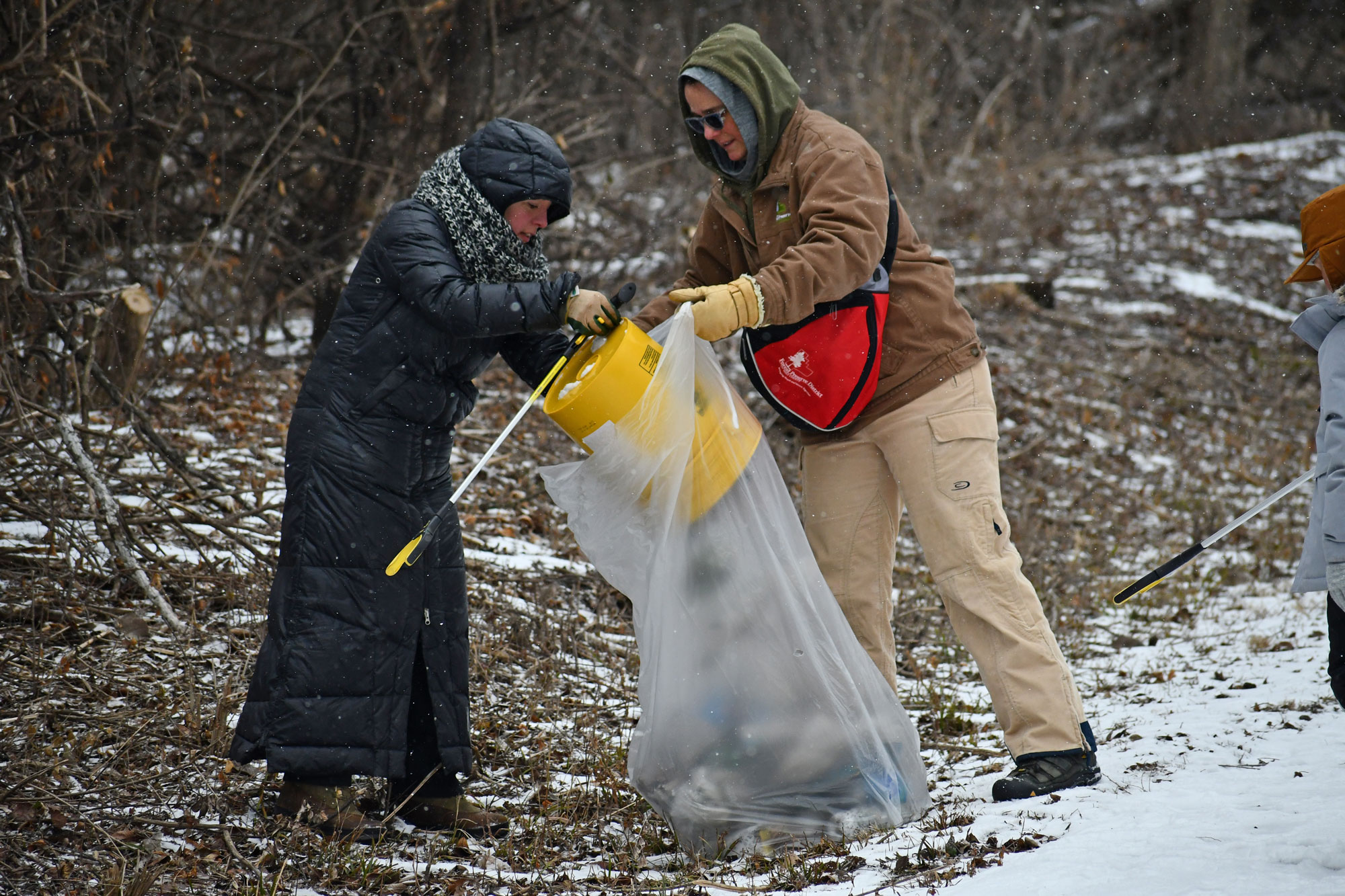 Two women pick up trash along a trail.