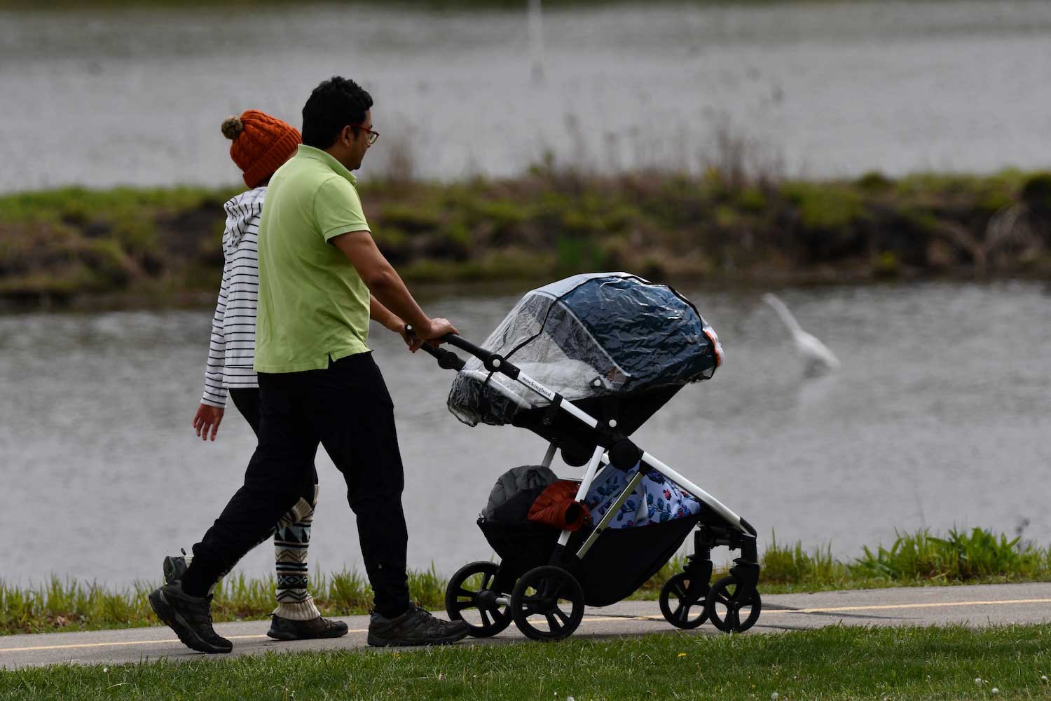 Two people, one pushing a stroller, walking along trail next to waterway.