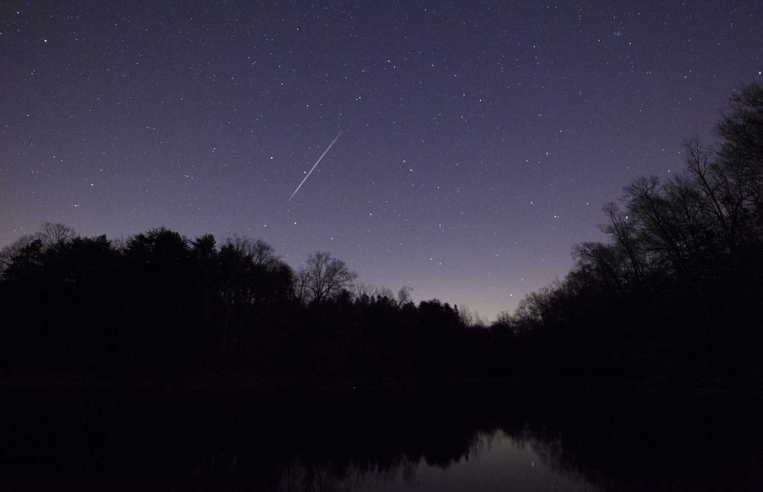 A meteor streaking across a star-filled night sky above trees and a lake.