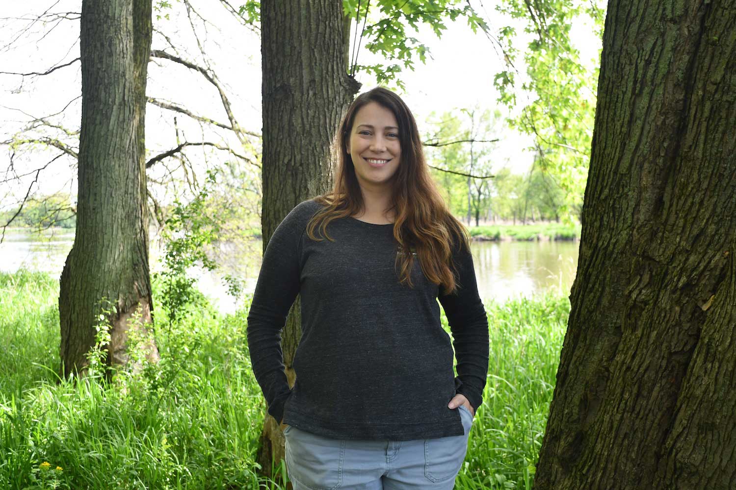 A person standing next to a tree with more trees and a river in the background.