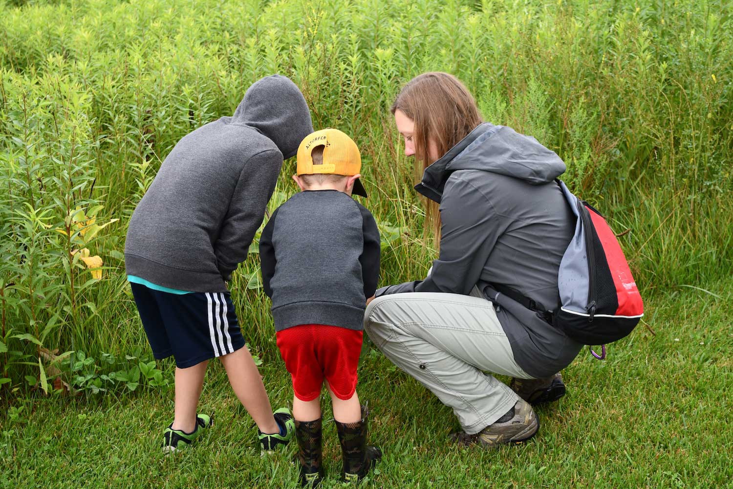 An adult and two young children crouched down in the grass looking at something with prairie plants in the background.