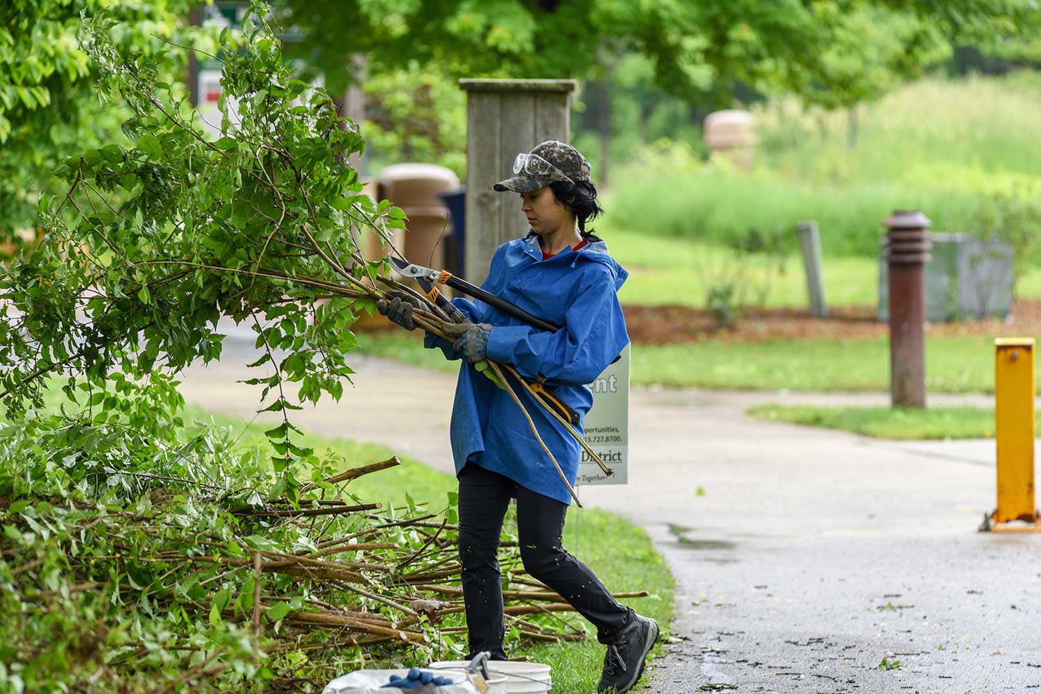 A person carrying loppers and cut brush while working alongside a paved trail.
