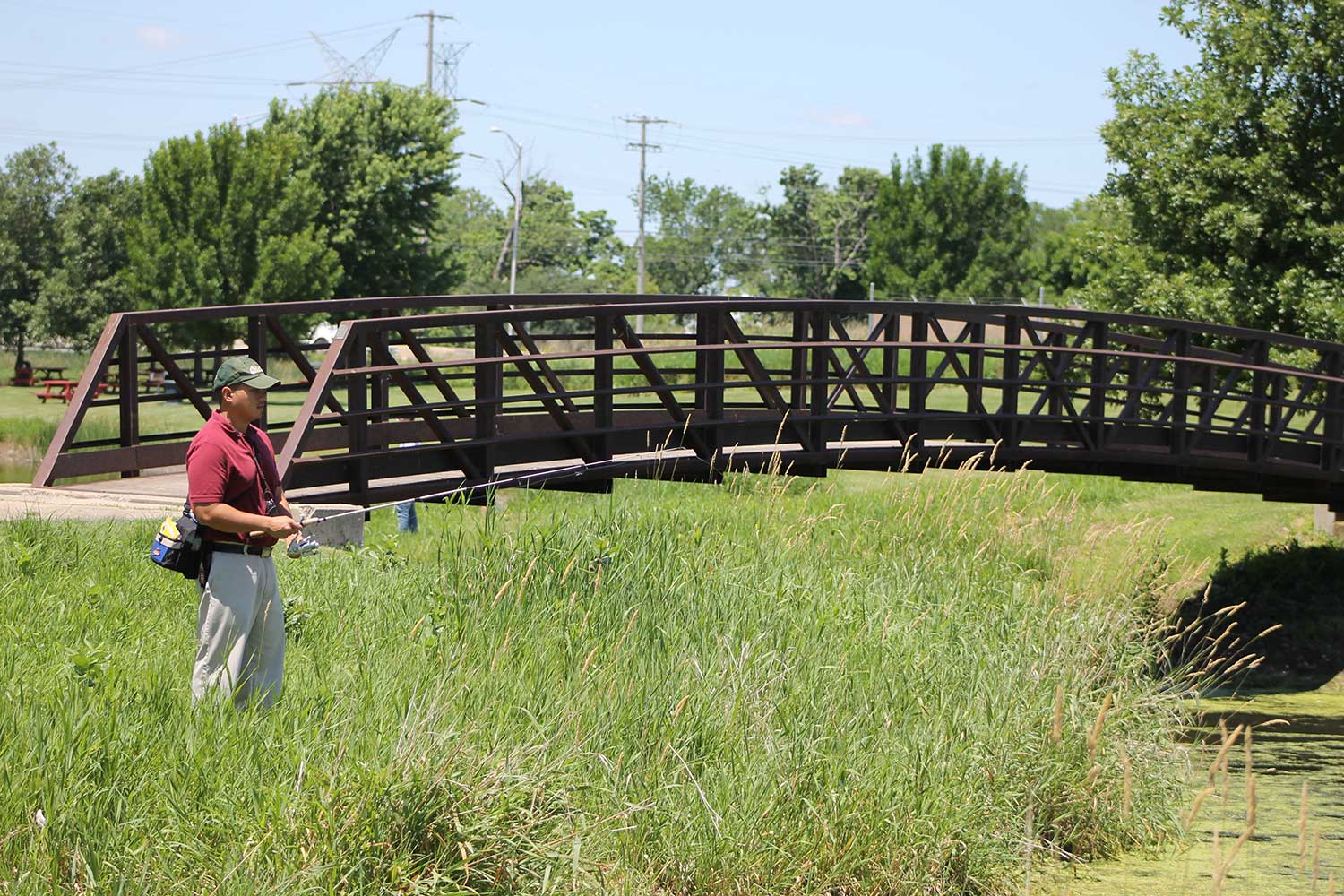 A person fishing along a shoreline with a footbridge in the background.