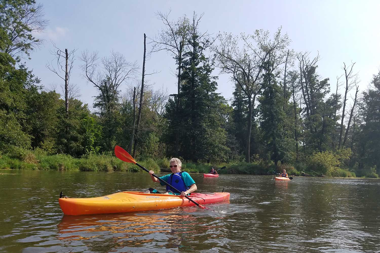 A person kayaking in a river with two more kayakers in the background.