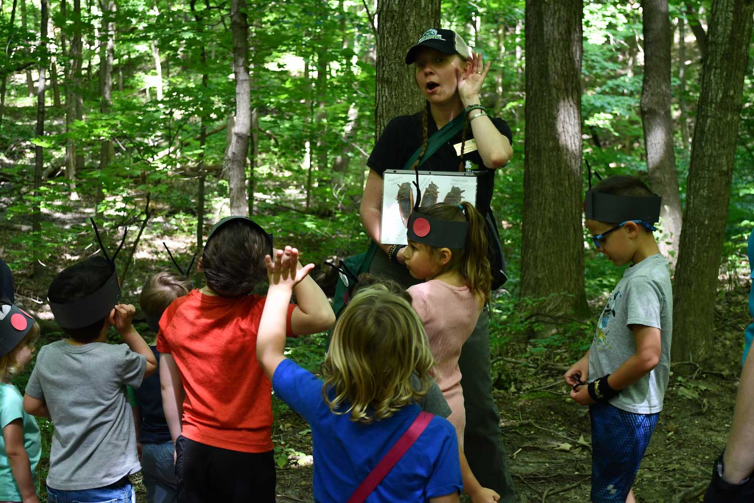 An adult standing in a forest with their hand to their ear as if listening while a group of children watch. 