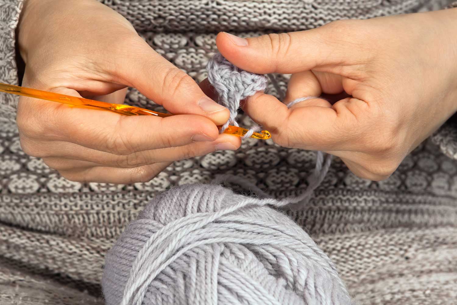 A closeup of a person holding crochet needles with a ball of yarn on a table.