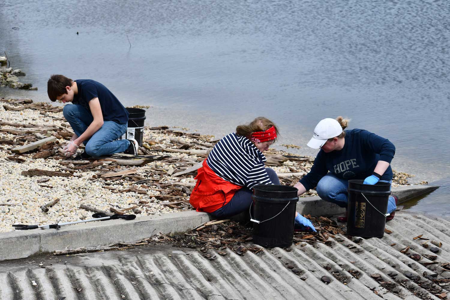 Three people crouched over on the ground picking up litter along a shoreline.