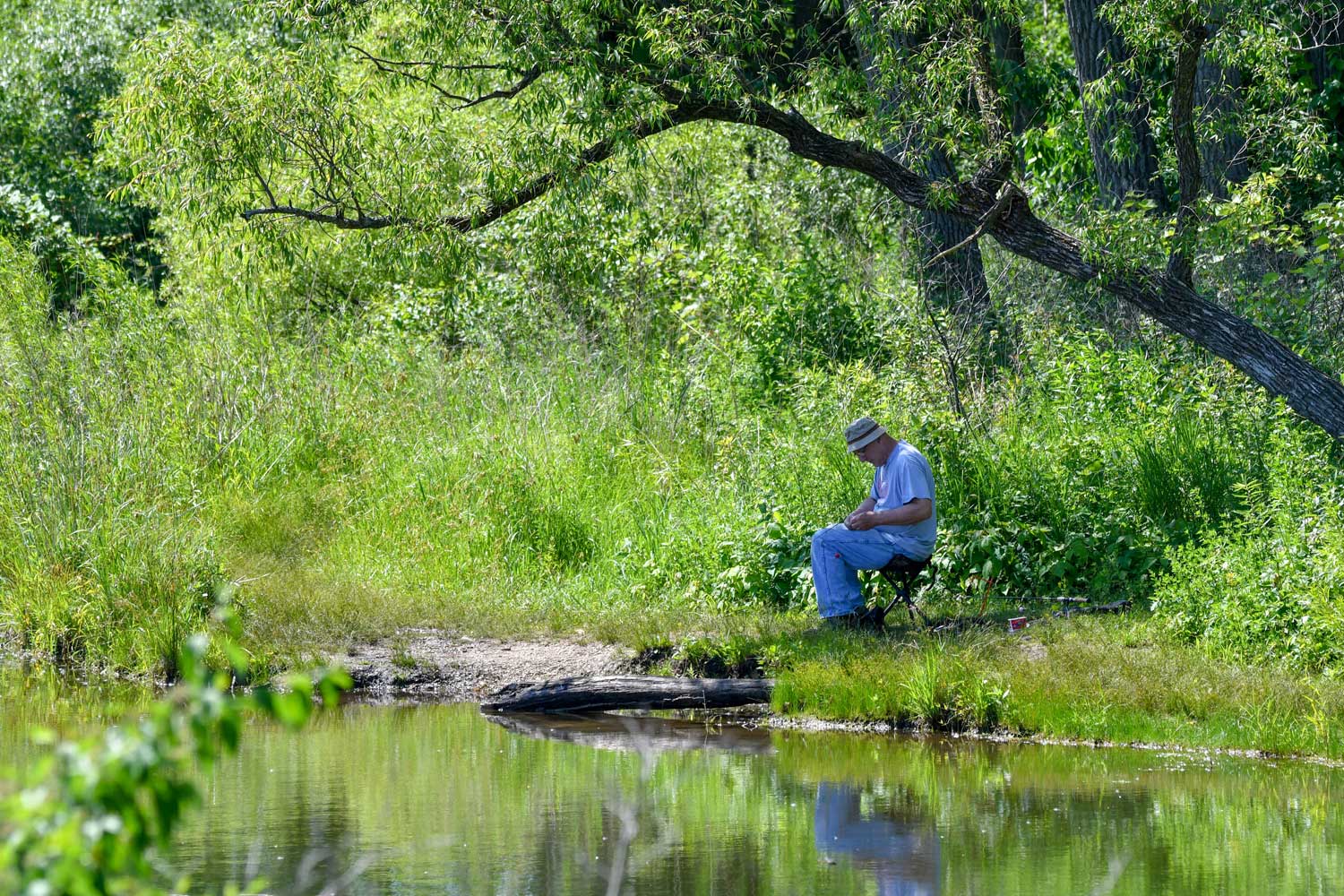 A man sitting along a shoreline getting a fishing pole ready to cast.