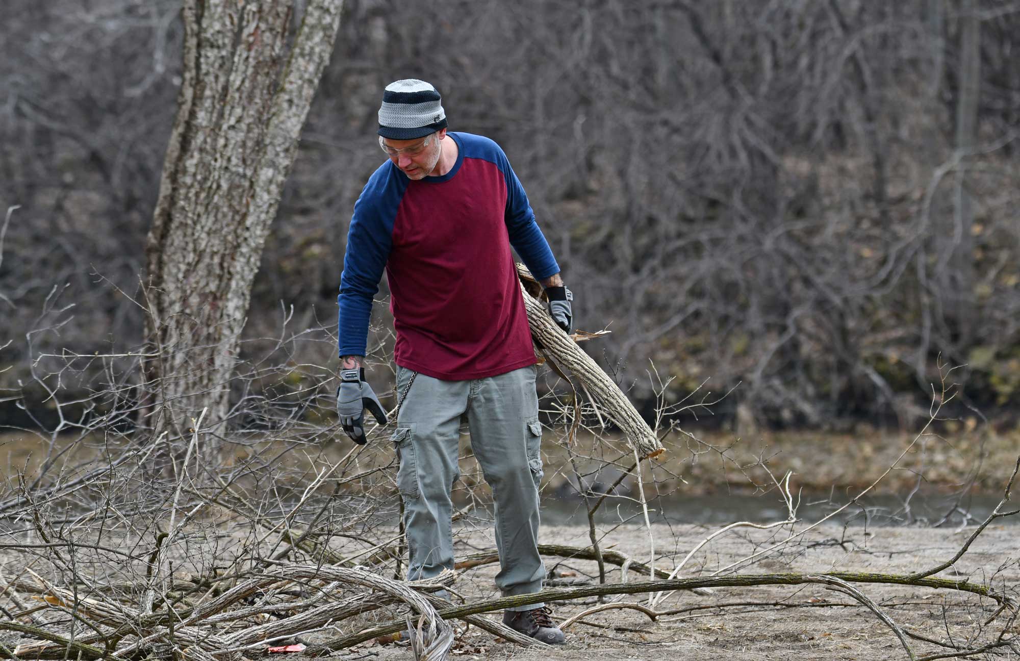 A person wearing gloves and googles carrying cut branches.