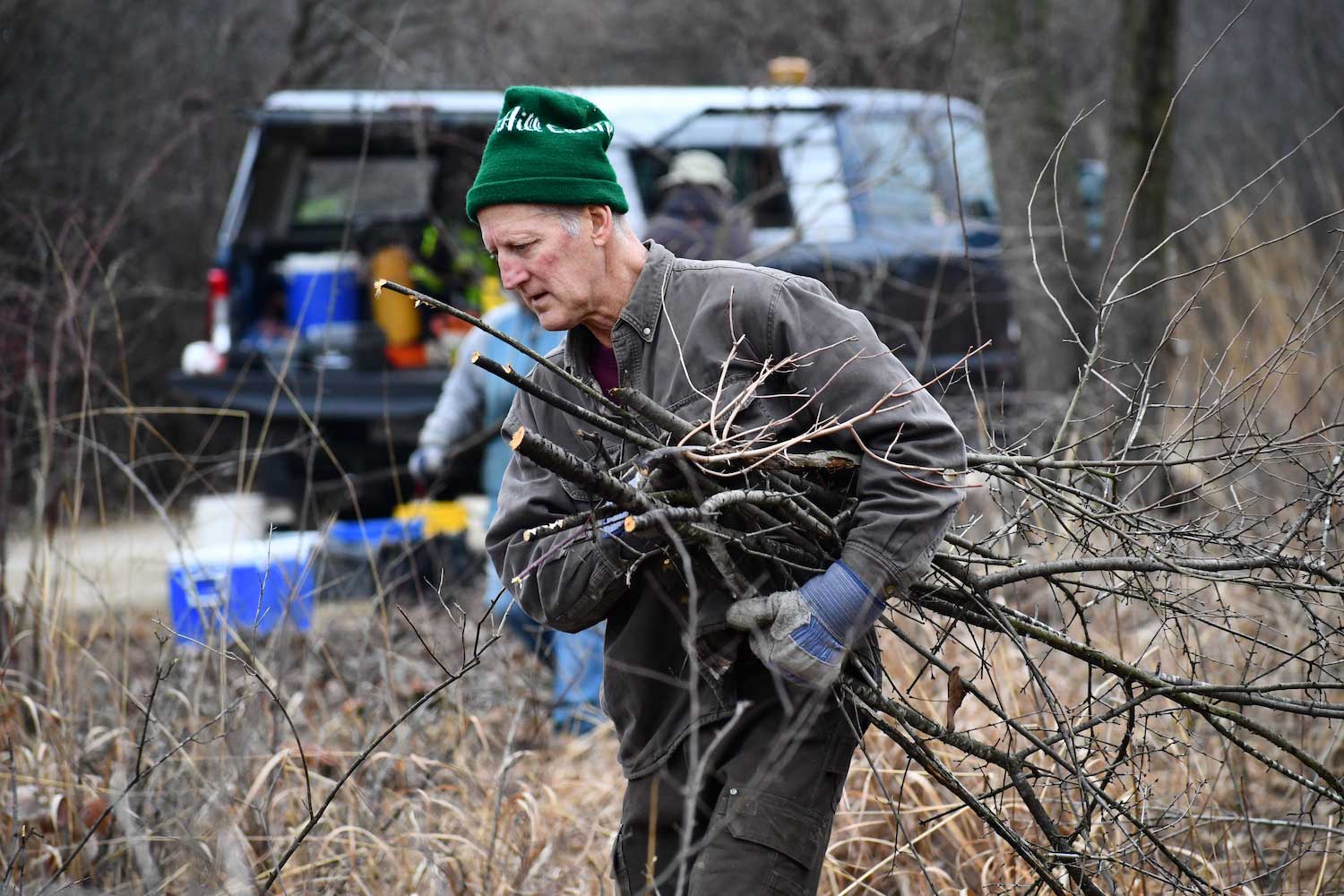 A person hauling branches across a natural area with a pickup truck full of equipment in the background.