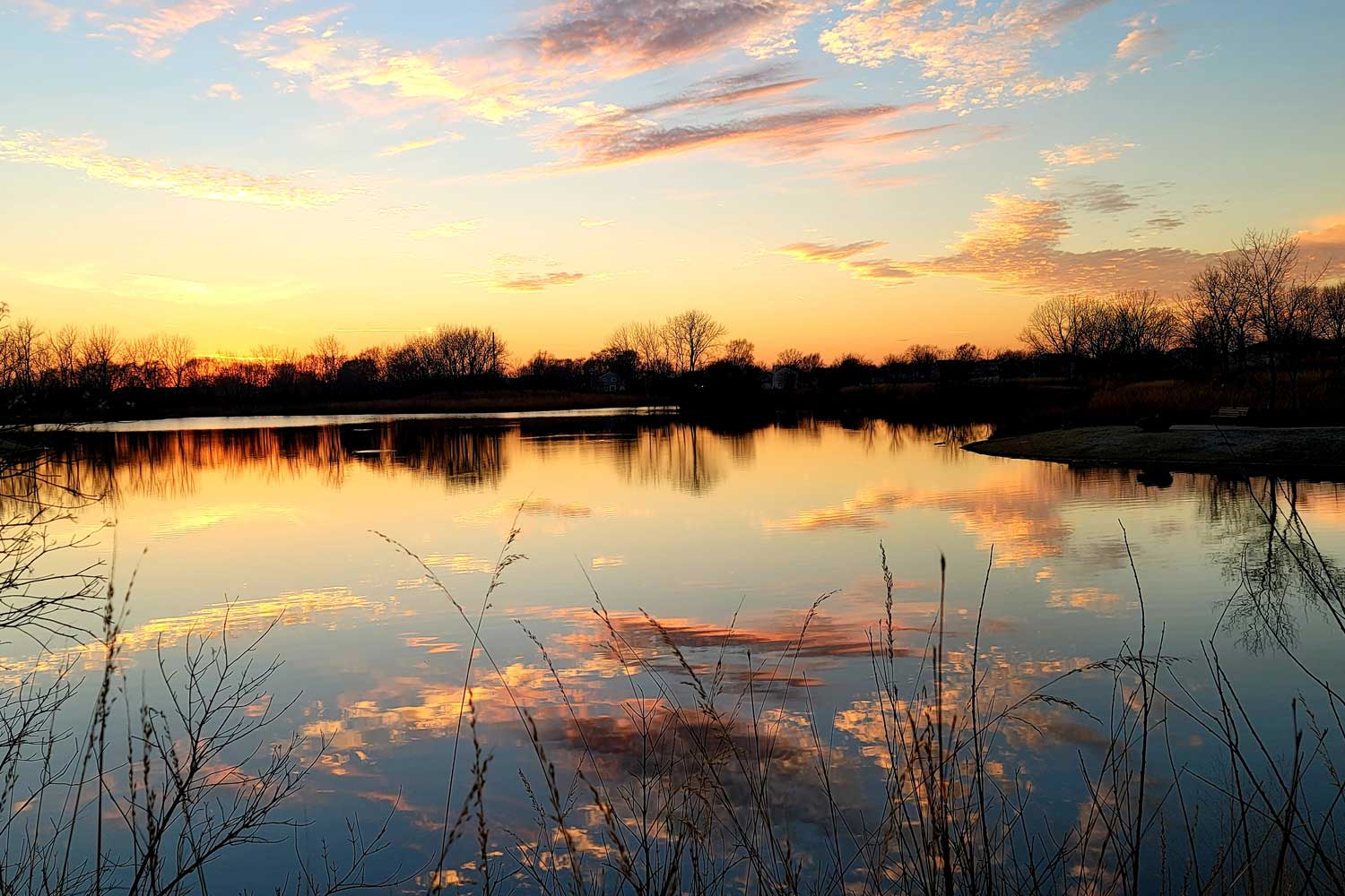 A colorful fall sunset with hues of yellow and orange reflected on the calm surface of a pond.