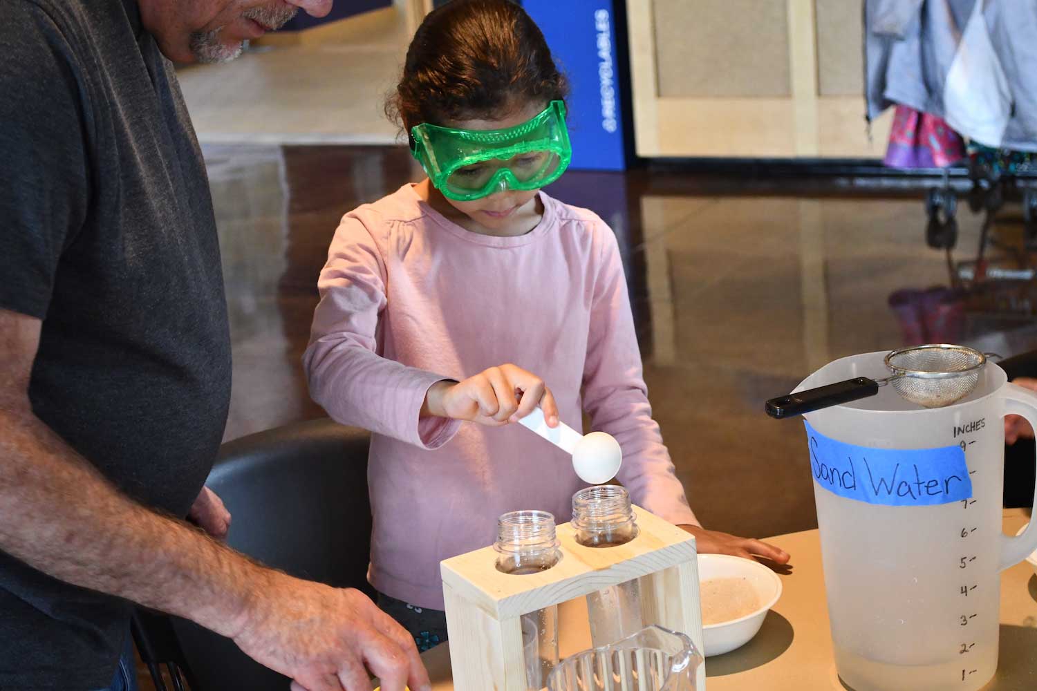 A child wearing goggles while standing at an table doing an experiment. 