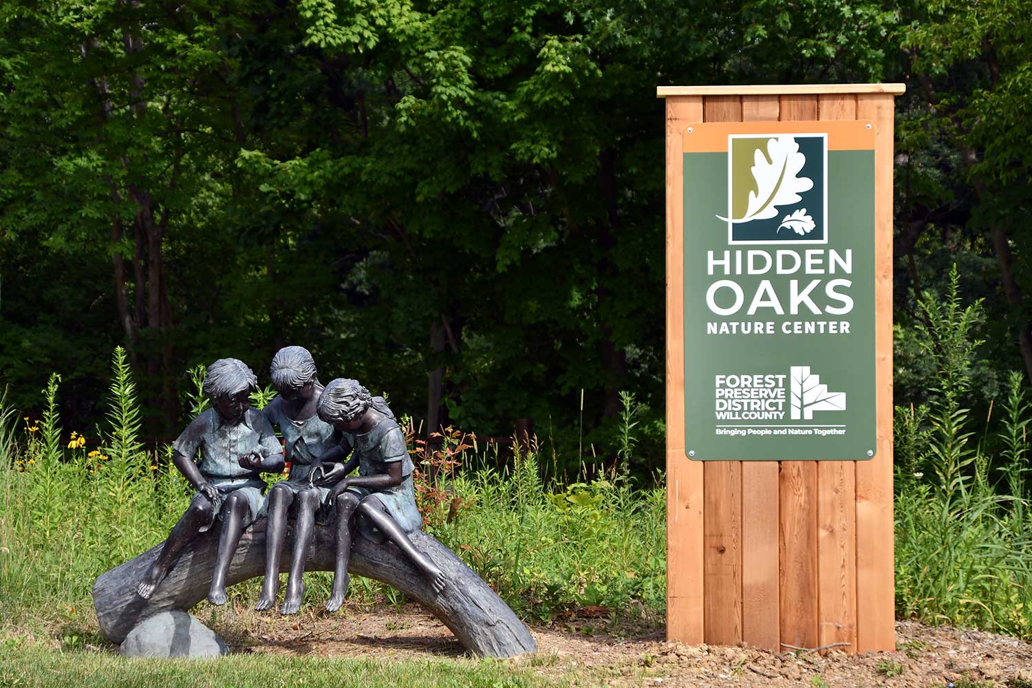 A Hidden Oaks sign and a structure of three children on a log.