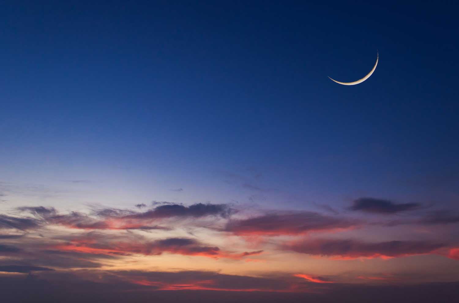 A new moon high in the sky above pinkish clouds just after the sun has set.