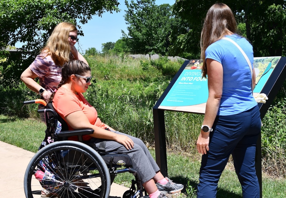 Three women on the all-abilities trail at Four Rivers: one in a wheelchair, one resting near the wheelchair and one explaining signage along the trail.
