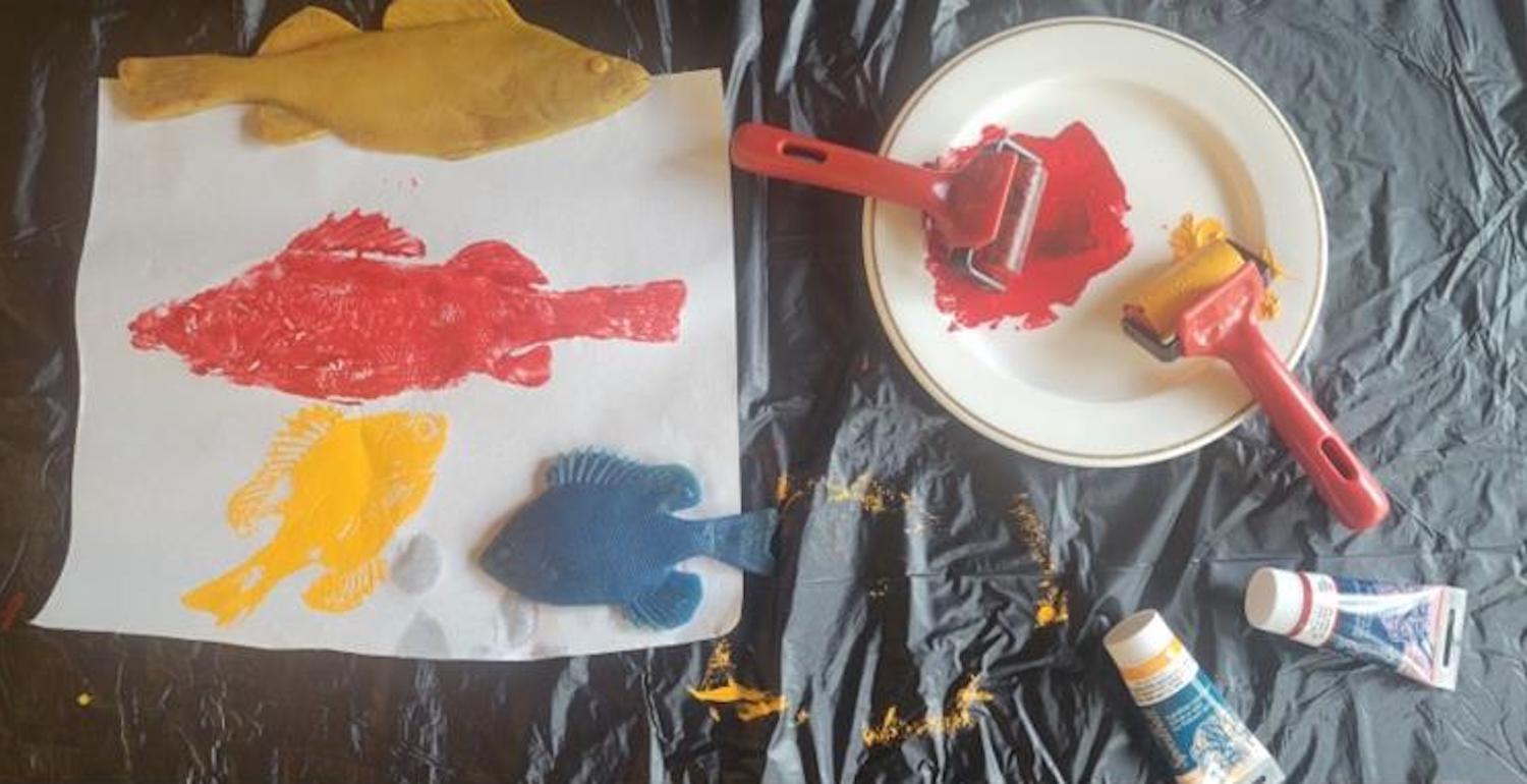 Fish models covered in yellow, red and blue paint next to fish prints on white paper.