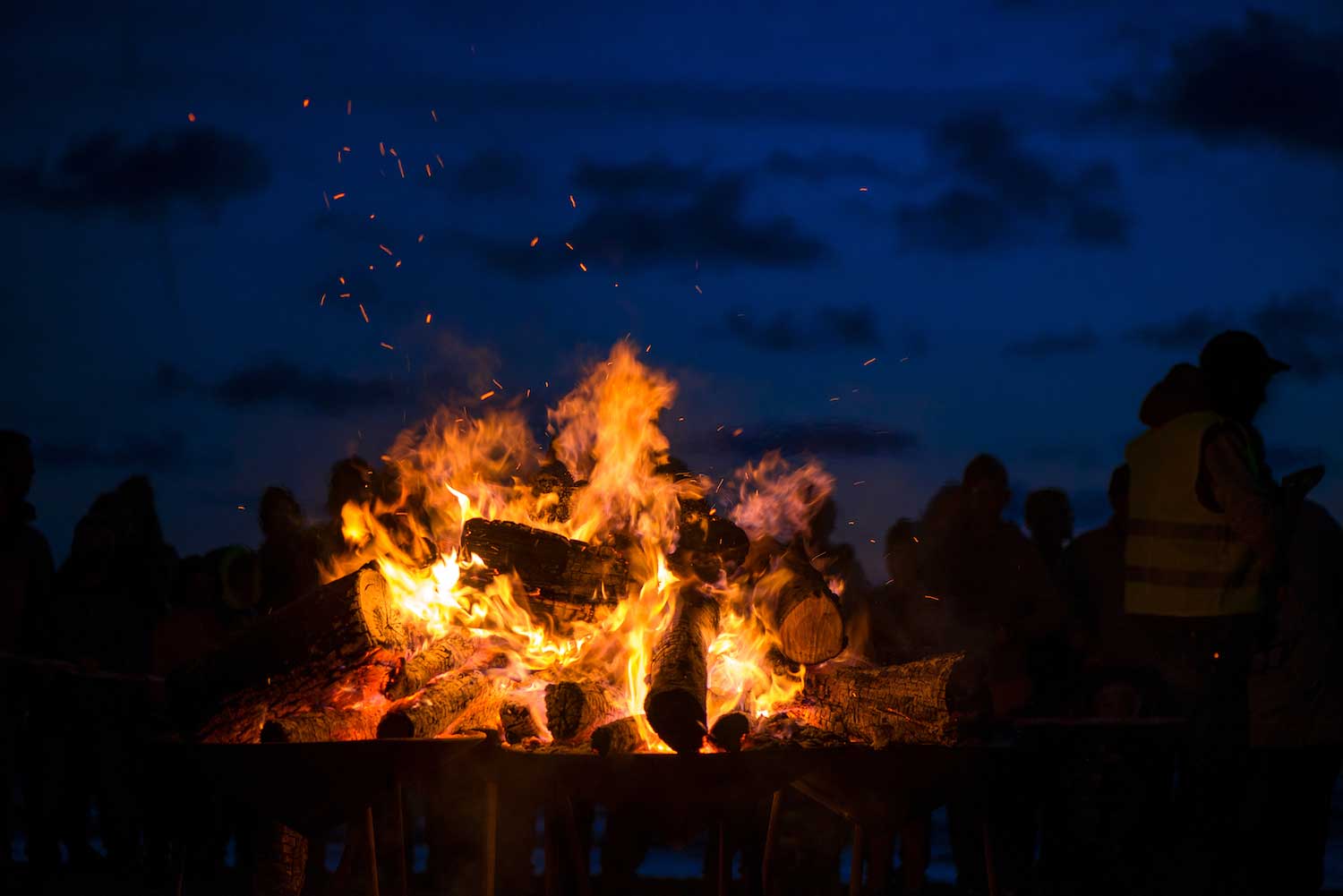 A bonfire burning in the dark with silhouettes faintly visible in the background.