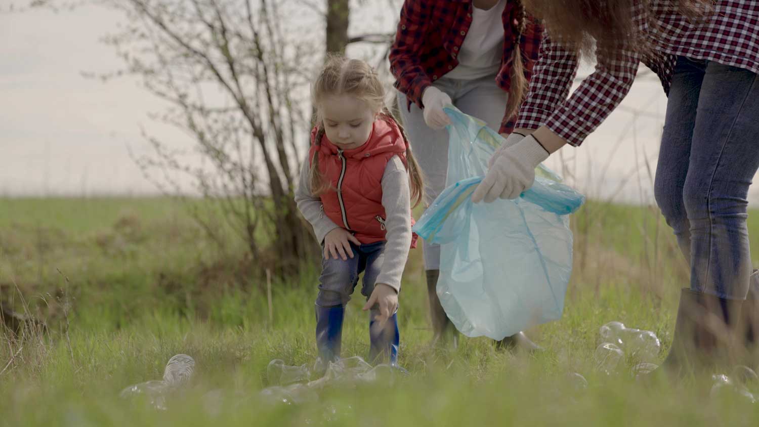 A child and two adults walking through a grassy area picking up trash.