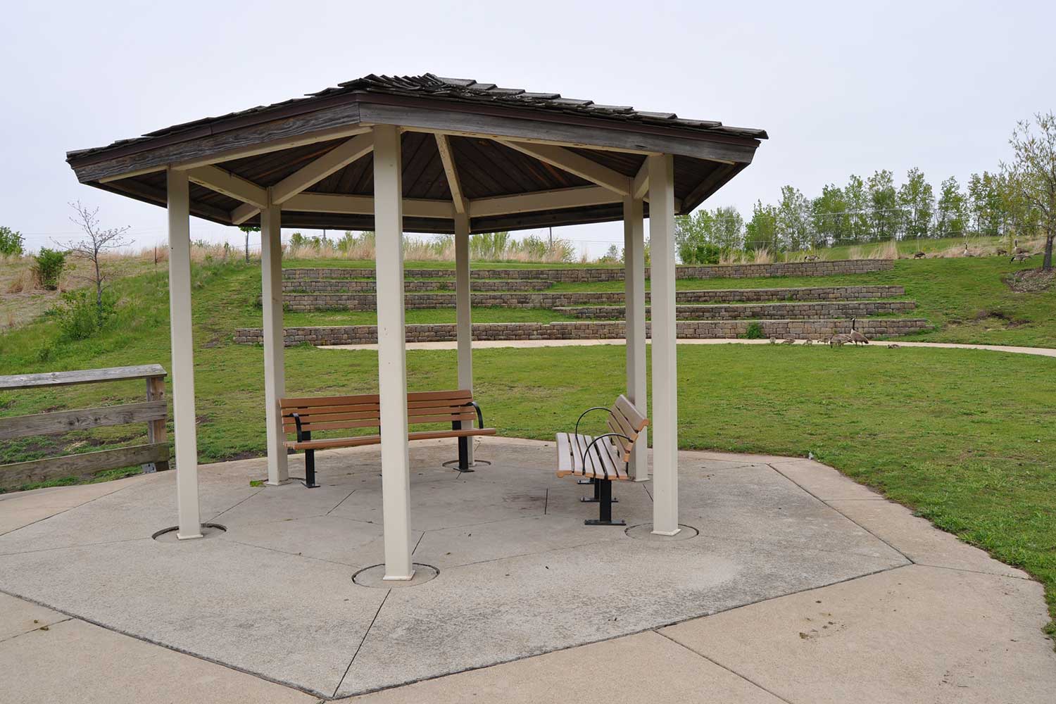 Two benches covered by a shade structure near a trail with grass in the background.