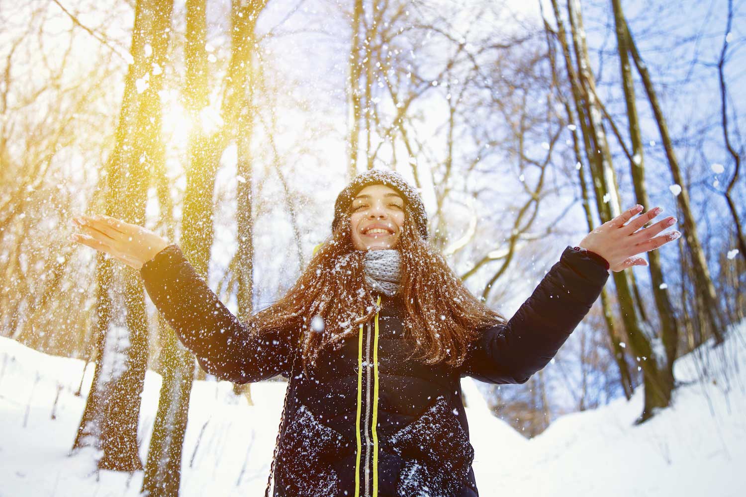 A person in winter gear standing with eyes closed and arms outstretched amidst a snowy winter scene with trees in the background.