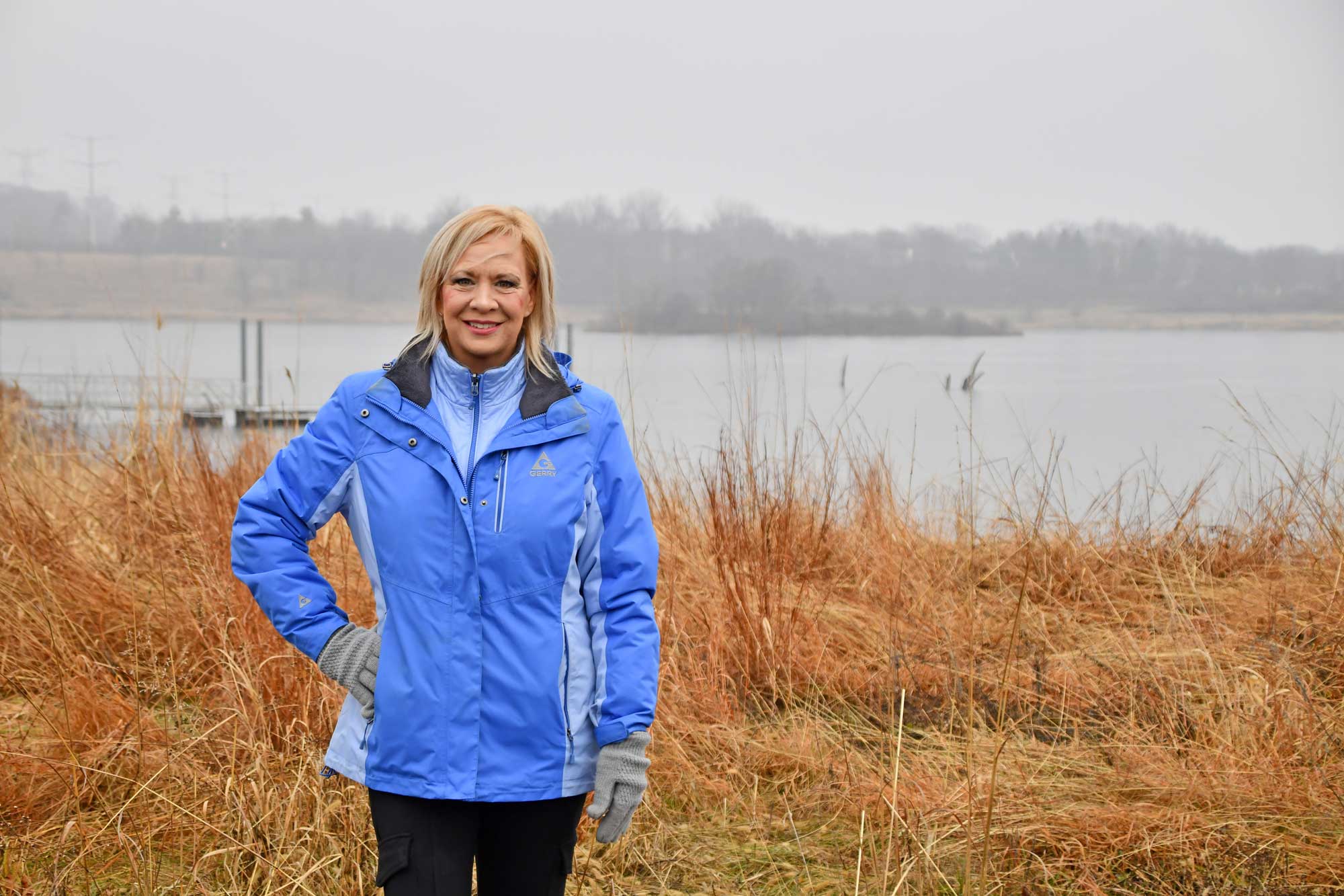 A woman in a blue jacket stands in a forest preserve with water in the background.