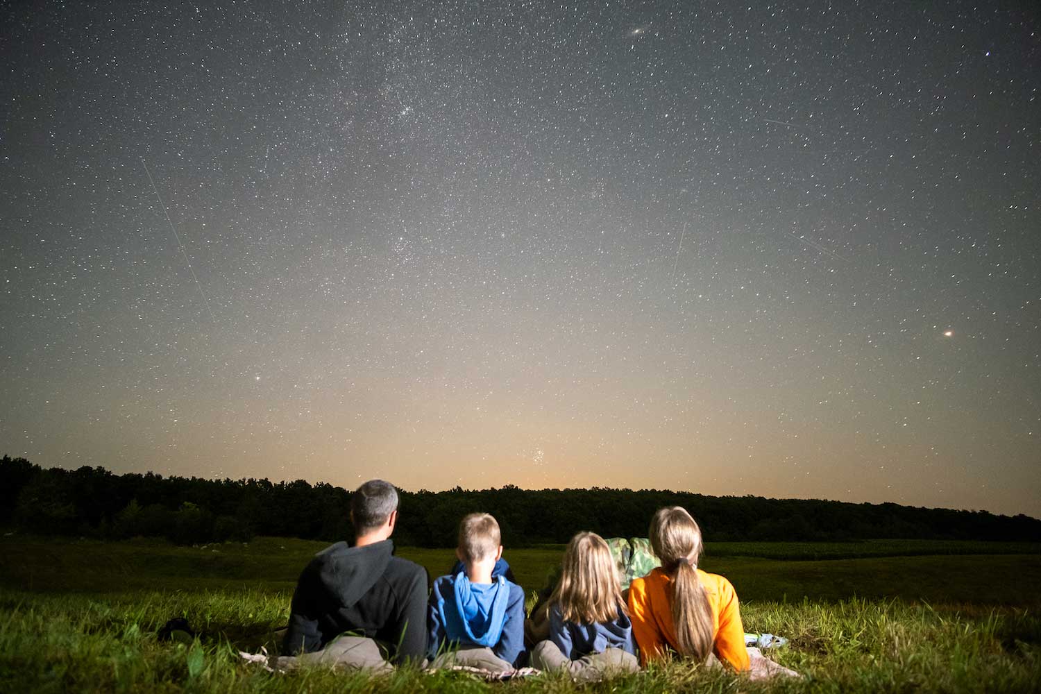 A group of two adults and two kids looking at the night sky while laying on a blanket in the grass.
