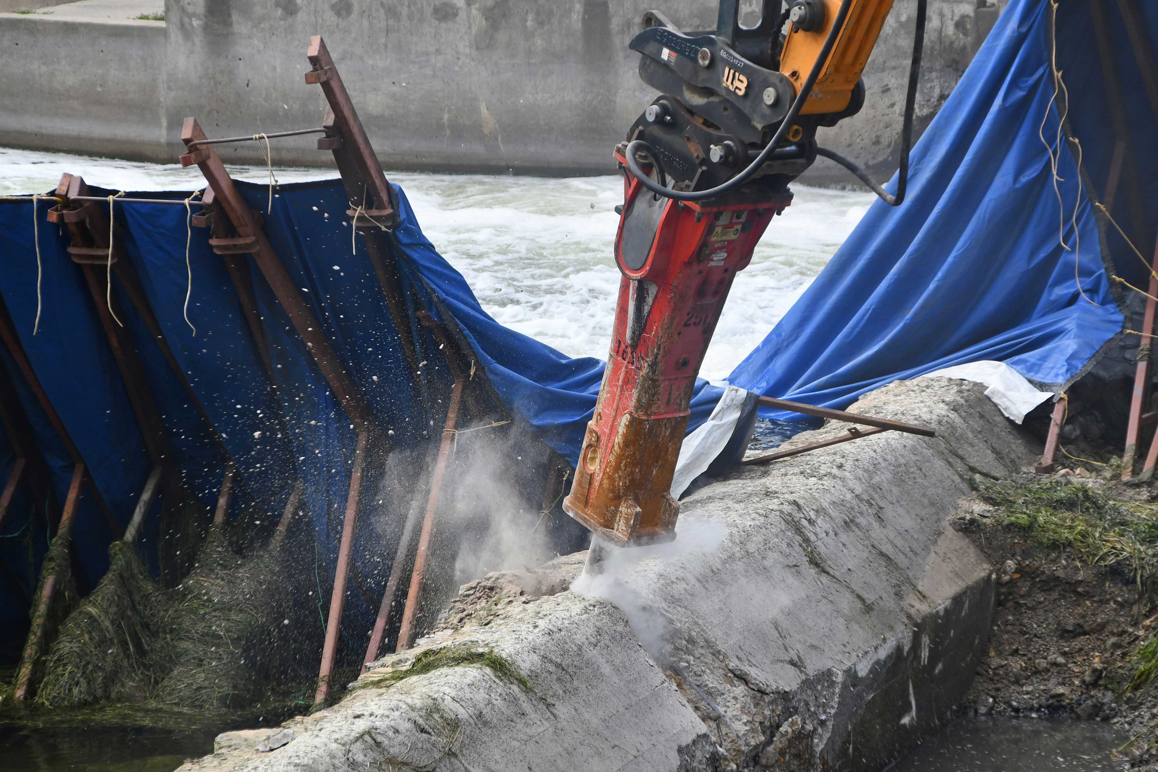 A hydraulic breaker on the end of an excavator chipping away at the dam. 