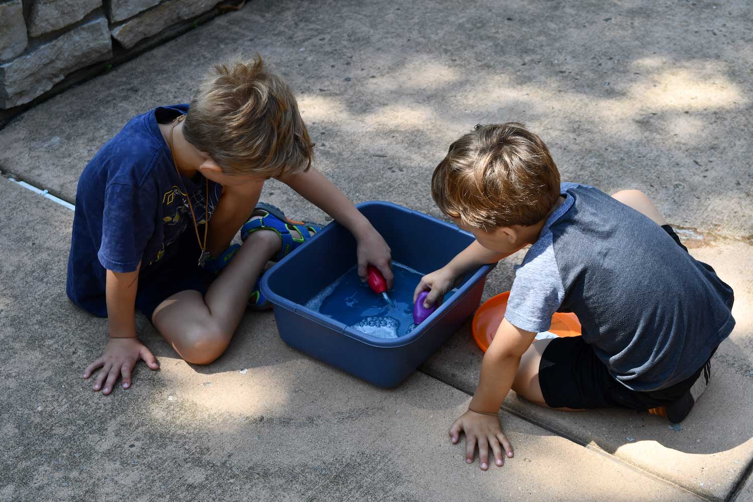Two your children sitting on a sidewalk playing with water in a tub.