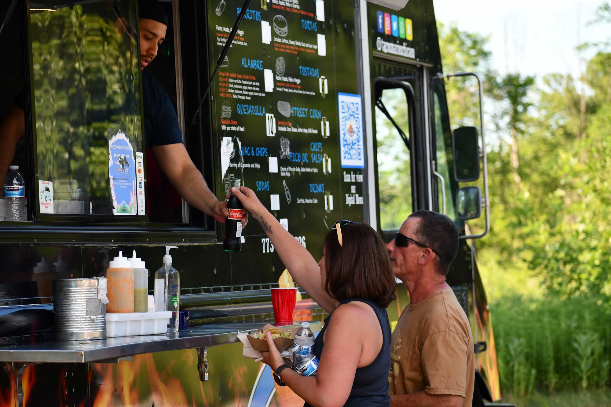 A man in a food truck hands a bottle of soda to a woman and man.